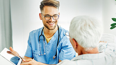 Fotografie eines fröhlich lachenden, blonden, jungen Altenpflegers mit Brille in blauem Arztkittel und mit Stethoskop um den Hals, der einer älteren Dame mit kurzen weißen Haaren, welche im Vordergrund von hinten zu sehen ist, nur eine Akte zeigt