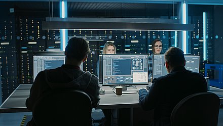 Vier Personen überwachen Computerbildschirme in einem dunklen Serverraum mit blauen Lichtern.