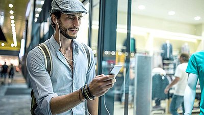 Herr mit Schiebermütze, Hemd, Rucksack und Kopfhörern im Ohr schaut vor Betreten eines Ladens auf sein Smartphone