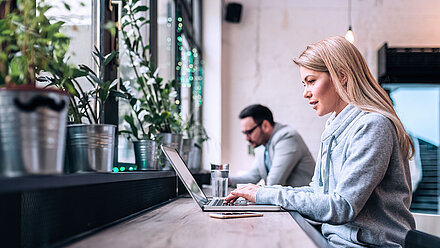 Foto von zwei Personen, die in einem modernen Büro oder Café an ihren Laptops arbeiten; links sind viele Topfpflanzen auf dem Fensterbrett gesehen; rechts davon sitzen eine junge, blonde Frau im Vordergrund und ein junger, brünetter Mann mit Brille im Hintergrund dem Fenster zugewandt an ihren Laptops und arbeiten gutgelaunt an etwas