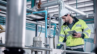 Foto: Techniker mit Schutzhelm, Warnjacke und Tablet steht zwischen Rohrleitungen und technischen Anlagenkomponenten in einer Industrieumgebung.