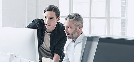 Junger IT-Mitarbeiter mit Zopf berät Kollegen mittleren Alters mit grauen Haaren und Bart zu Sicherheitseinstellungen am PC