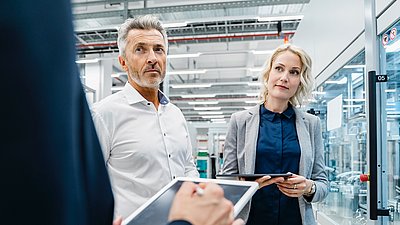 Foto: Zwei Personen stehen mit Tablets in einer modernen Industrie- oder Produktionsumgebung mit verglasten Maschinenbereichen und technischen Anlagen.