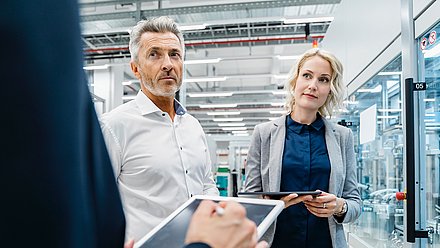 Foto: Zwei Personen stehen mit Tablets in einer modernen Industrie- oder Produktionsumgebung mit verglasten Maschinenbereichen und technischen Anlagen.