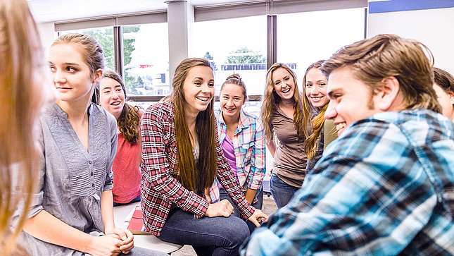 Gruppe lächelnder Schülerinnen und Schüler sitzt zusammen in einem hellen Klassenraum und unterhält sich.