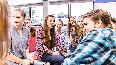 Gruppe lächelnder Schülerinnen und Schüler sitzt zusammen in einem hellen Klassenraum und unterhält sich.