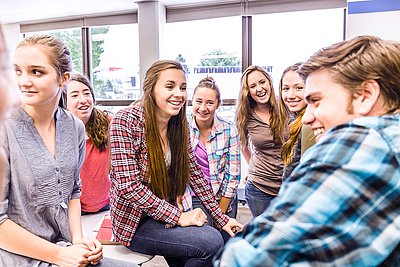 Gruppe lächelnder Schülerinnen und Schüler sitzt zusammen in einem hellen Klassenraum und unterhält sich.