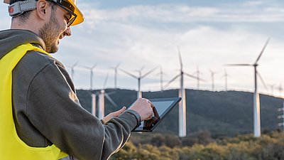 Foto: Person mit Schutzhelm und Warnweste bedient ein Tablet vor mehreren Windkraftanlagen in einer Hügellandschaft.