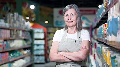 Filialleiterin mittleren Alters mit grauen langen Haaren und Arbeitsschürze steht lächelnd zwischen zwei Supermarktregalen und lächelt zufrieden