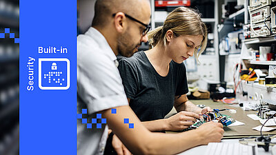 Zwei Personen arbeiten gemeinsam an Elektronikbauteilen in einer Werkstatt, LANCOM Built-in Security links eingeblendet.
