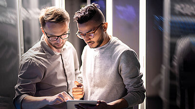 Zwei Männer stehen in einem Serverraum und analysieren gemeinsam Daten auf einem Tablet.