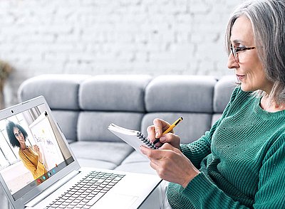 Ältere Dame mit grauen Haaren, Brille und grünem Pullover sitzt an Couchtisch mit Stift und Zettel vor Laptop und notiert sich wichtige Informationen aus einem Webinar