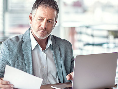 Mann mittleren Alters mit grauen Haaren und Bart sitzt nachdenklich vor Laptop und betrachtet ein Stück Papier