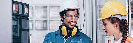 Black-haired worker in utility plant with white hard hat, safety goggles and yellow noise protection headphones in blue overalls and work gloves holding laptop and standing to the right of a control box (left); he explains something to his brunette colleague to his right, smiling; his colleague is also wearing a yellow hard hat and safety vest and listening attentively