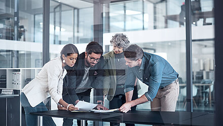 Zwei Frauen und zwei Männer beugend sich stehend über einen Tisch und sehen sich gemeinsam verschiedene Dokumente an, die auf dem Tisch verteilt sind.