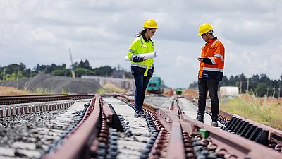 Foto: Zwei Personen mit Schutzhelmen und Warnkleidung stehen auf Bahngleisen und prüfen Unterlagen beziehungsweise ein Tablet.