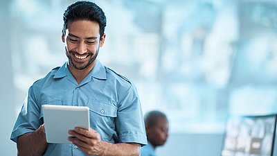 Foto: Sicherheitsmitarbeiter in blauer Arbeitskleidung nutzt ein Tablet zur mobilen Steuerung und Überwachung integrierter Sicherheits- und Partnerlösungen in einer modernen KRITIS-Umgebung.