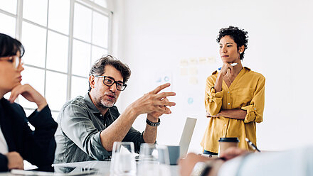 Im Fokus ist ein Mann sitzend an einem Tisch, der gestikulierend etwas erklärt. Daneben steht eine Frau mit einem Stift in der Hand, die interessiert zuhört.
