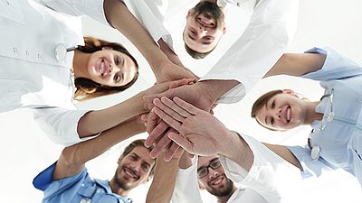 Medizinisches Team legt Hände übereinander und lächelt, Symbol für Zusammenarbeit und Teamgeist im Gesundheitswesen.