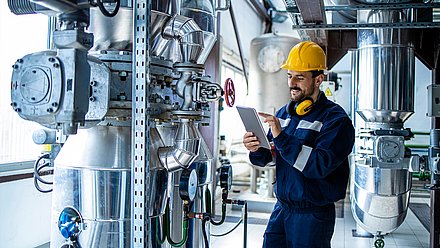 Foto: Techniker mit Schutzhelm und Tablet steht zwischen industriellen Rohrleitungen, Ventilen und technischen Anlagenkomponenten.
