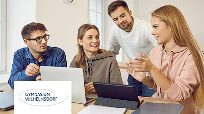 Vier Schüler diskutieren lebhaft an Laptops und Tablets in heller Lernumgebung. In Anlehnung an das Gymnasium Wilhelmsdorf.