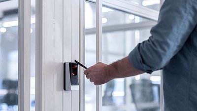 Foto: Person nutzt ein Smartphone an einem elektronischen Zutrittsleser zur sicheren Zugangskontrolle in einem modernen Gebäude und veranschaulicht digitale Sicherheits- und Zutrittskonzepte für kritische Infrastrukturen.