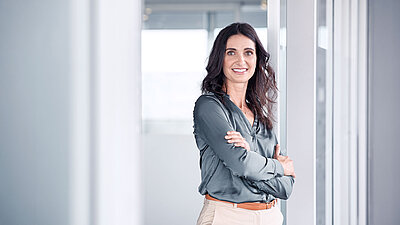 Frau lehnt mit verschränkten Armen an einer Glastür und lächelt in die Kamera