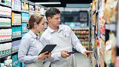 Eine Supermarktmitarbeiterin mit Tablet prüft mit ihrem Kollegen mit Handheld-Scanner die Ware im Regal