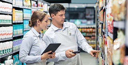 Eine Supermarktmitarbeiterin mit Tablet prüft mit ihrem Kollegen mit Handheld-Scanner die Ware im Regal