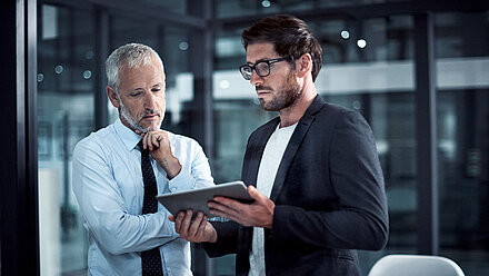 Ein Mann hält ein Tablet in der Hand und hält es einem anderen Mann hin, sodass Sie beide auf den Bildschirm gucken können.