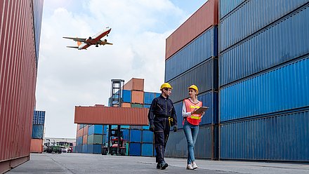 Foto: Zwei Personen mit Schutzhelmen laufen zwischen hohen Containerstapeln eines Containerterminals; im Hintergrund fährt ein Containerstapler, darüber fliegt ein Passagierflugzeug.