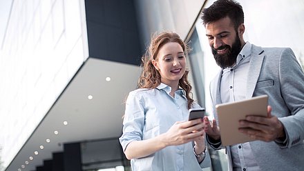 Foto: Zwei Personen stehen vor einem modernen Gebäude und schauen gemeinsam auf Smartphone und Tablet.