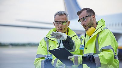 Foto: Zwei Mitarbeitende in gelber Schutzkleidung und mit Gehörschutz stehen vor einem Flugzeug und besprechen Unterlagen auf einem Klemmbrett.