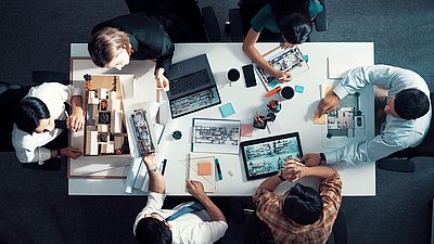 Foto: Mehrere Personen sitzen an einem Tisch mit Architekturmodellen, Grundrissen, Tablets und Laptop und arbeiten gemeinsam an der Planung eines Gebäudesystems.