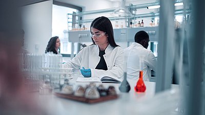 Foto: Mehrere Personen arbeiten in einem modernen Labor; im Vordergrund sitzt eine Frau mit Schutzbrille und Handschuhen an einem Labortisch mit Glasgefäßen und Proben
