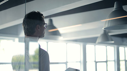 Mann steht mit einem Tablet in der Hand hinter einer Glaswand eines Büros