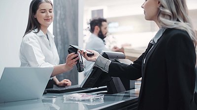 Blonde Kundin in schwarzem Blazer bezahlt kontaktlos mit ihrem Smartphone an der Kasse eines modernen Geschäfts
