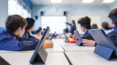 Schüler sitzen mit Tablets im Klassenzimmer und folgen dem Unterricht, Lehrkraft zeigt auf das Smartboard.