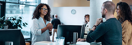 Verwaltungsmitarbeiterin mit schwarzen Haaren und Brille begrüßt freundlich junges Paar an ihrem digitalen Arbeitsplatz