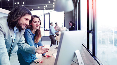 Frau und Mann lachend vor PC-Bildschirm in modernem Büro