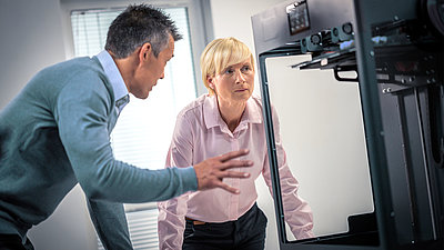 Blonde Frau mittleren Alters mit kurzen Haaren diskutiert vor einer Maschine angeregt mit ihrem männlichen Kollegen