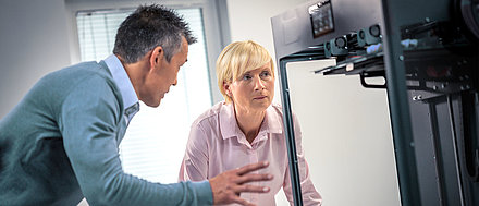 Blonde Frau mittleren Alters mit kurzen Haaren diskutiert vor einer Maschine angeregt mit ihrem männlichen Kollegen