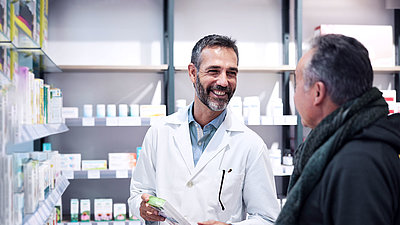 Zwei Männer in einer Apotheke; Apotheker mit kurzen, schwarzen Haare und Bart; trägt einen Arztkittel und hält ein Medikament in der Hand. Er lächelt einen Kunden an, der vor ihm steht, auch schwarze, kurze Haare, Jacke und Schal.