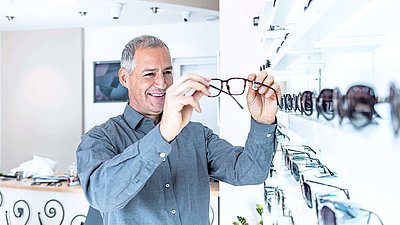 Älterer Mann mit grauen Haaren steht bei Optiker vor Brillenregal und betrachtet zufrieden eine Brille