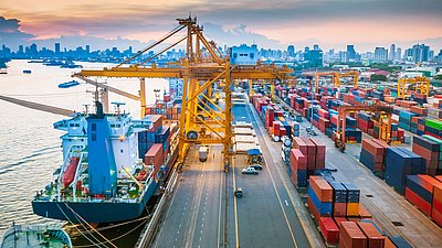 Foto: Großer Containerhafen mit Frachtschiff, Kränen und zahlreichen Containerstapeln vor einer Stadtsilhouette.