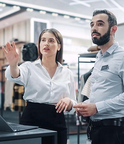 Brünette Filialleiterin in weißer Bluse zeigt aufmerksamen Angestelltem wo das nächste digitale Werbebanner in einem Modeladen platziert wird