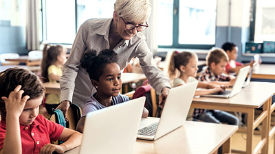 Blonde Lehrerin mit Brille prüft digitale Aufgabe, die eine Grundschülerin am Laptop gelöst hat, während jedes Kind im Klassenzimmer an Laptop arbeitet