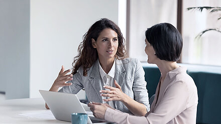 Zwei Kolleginnen sitzen am Tisch vor einem Laptop, eine gestikuliert mit ihren Händen