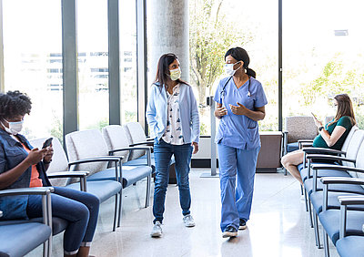 Junge Ärztin in hellblauem Overall holt Patientin mit Mundschutz aus hellem Wartebereich einer Klinik für ihre Behandlung ab, wo eine Schwangere und eine weitere Frau warten, dass sie aufgerufen werden