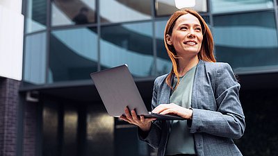 Foto: Frau mit Laptop steht vor einem modernen Bürogebäude mit Glasfassade.